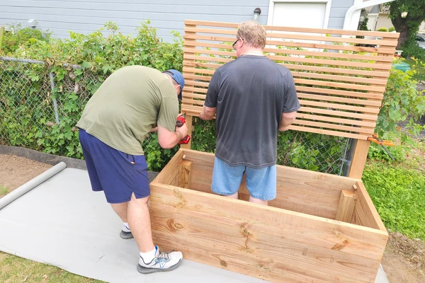 view of a garden bed with a privacy panel in the process of being built by two men currently screwing in the wood slats to the vertical supports.