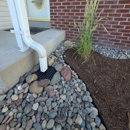 drainage pipe from house leading into catch basin, surrounded by river rock with larger rock detailing the basin for added appeal to landscape design